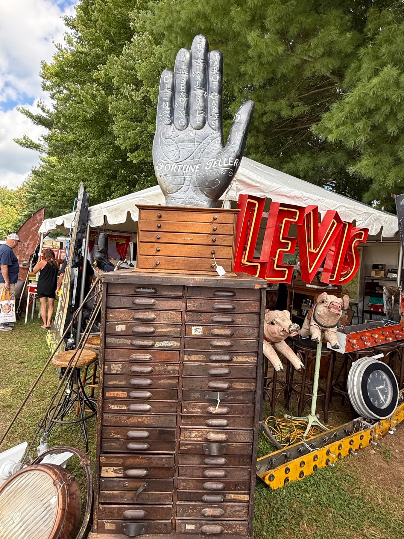 A vintage dealer booth at Brimfield with a Fortune Teller hand sign, a Levi's neon sign, and a wall of flat file cabinets